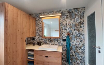 Badezimmer mit Holzschrank, offenem Regal mit Handtüchern, weißem Waschbecken, Spiegelschrank mit Beleuchtung und türkisfarbenem Handtuch an einem Wandhaken.