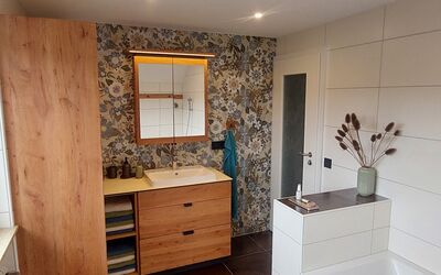 Helles Badezimmer mit großem Fliesenboden, Holzschränken, weißer Badewanne, dekorativer Vase mit Trockenblumen und Waschbecken mit beleuchtetem Spiegelschrank vor floral gemusterter Wand.